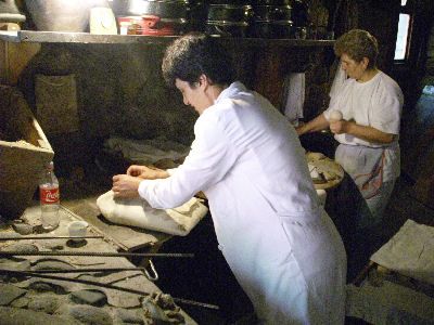 Een deegstukje wordt op een soort ovenwant geplaatst, achterin de bakkerij worden deegstukken opgebold.