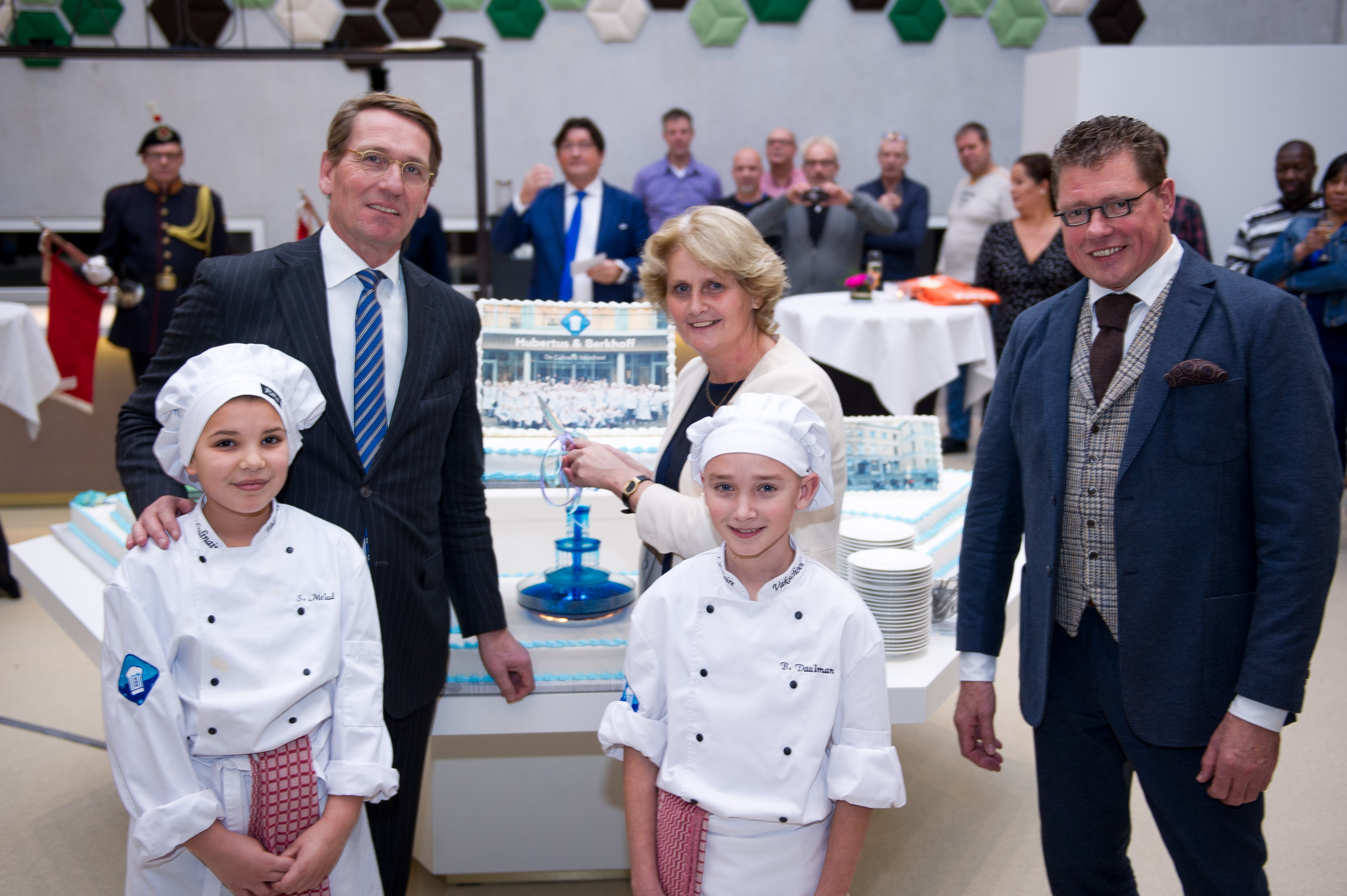 Edo de Jaeger (voorzitter van de raad van bestuur), Nel Winkel (directeur afdeling Onderwijs gemeente Amsterdam), Ruud Versteeg (schooldirecteur Hubertus & Berkhoff) (v.l.n.r.) en twee leerlingen snijden de feestelijke taart aan. Foto: Martijn Buskermolen
